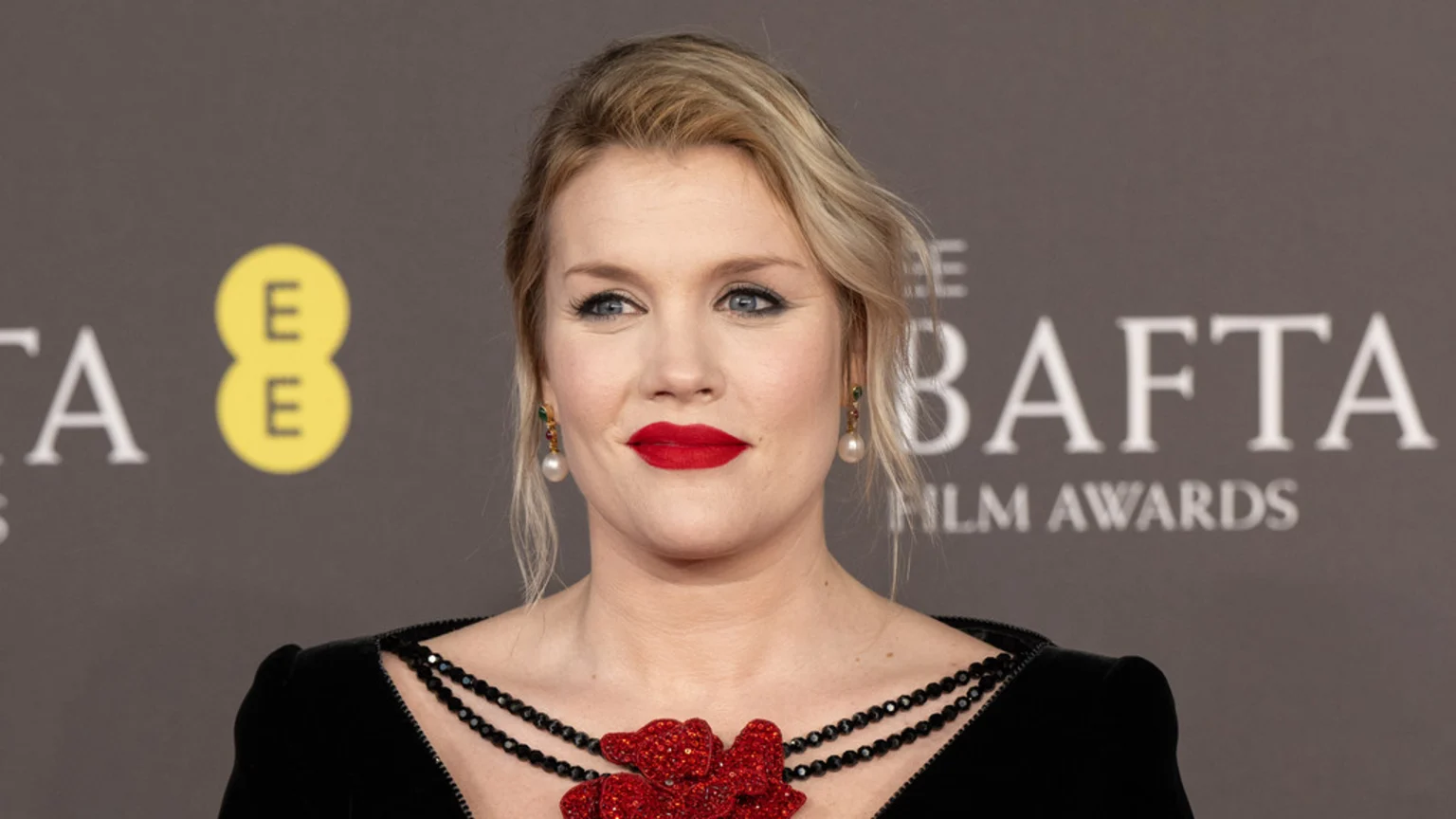 A blonde woman with red lipstick and winged eyeliner wears a black dress with a red beaded flower on the chest and pearl drop earrings, posing against a grey BAFTA background.