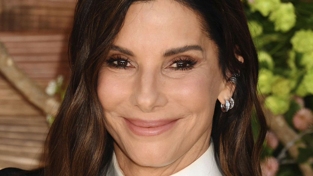A close-up portrait of actress Sandra Bullock, with long dark hair and a subtle smile, wearing a white collared shirt and silver hoop earrings, against a blurred background.