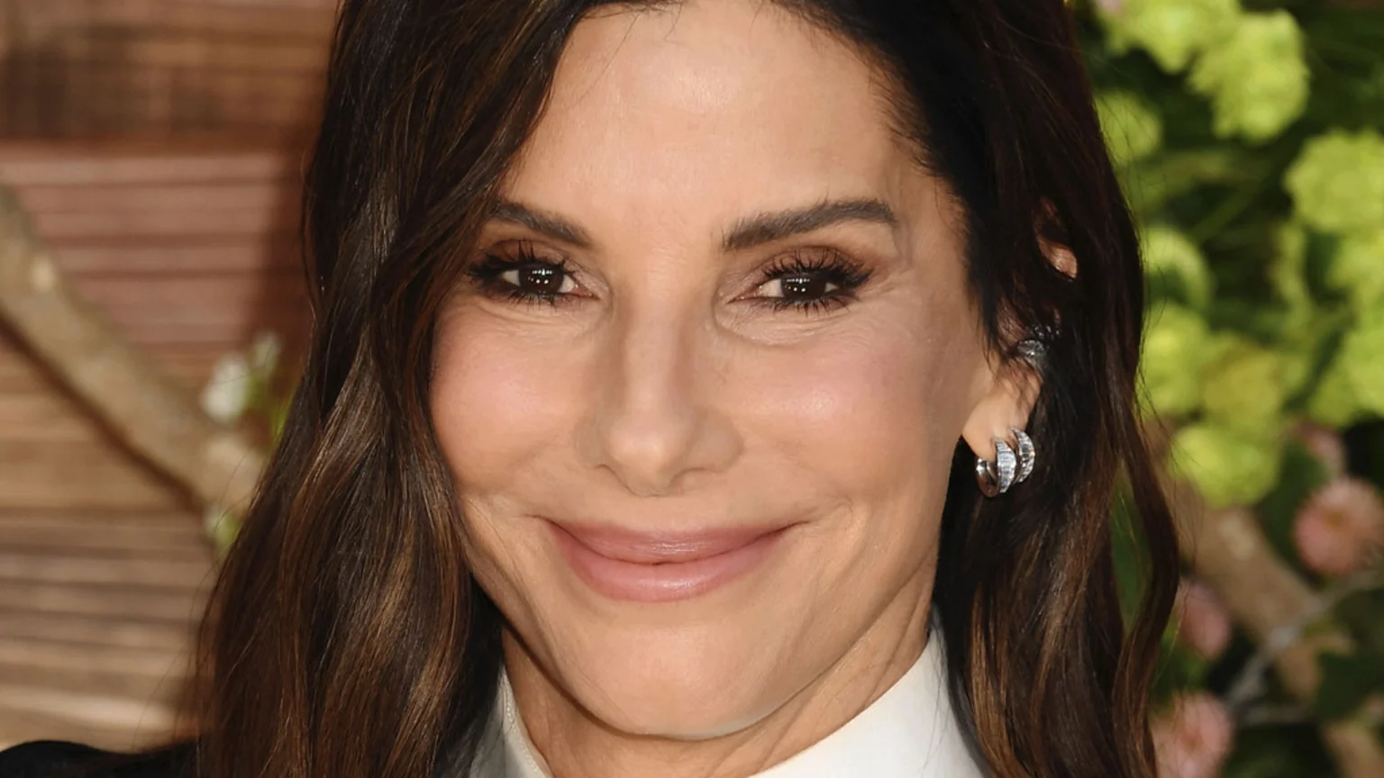 A close-up portrait of actress Sandra Bullock, with long dark hair and a subtle smile, wearing a white collared shirt and silver hoop earrings, against a blurred background.