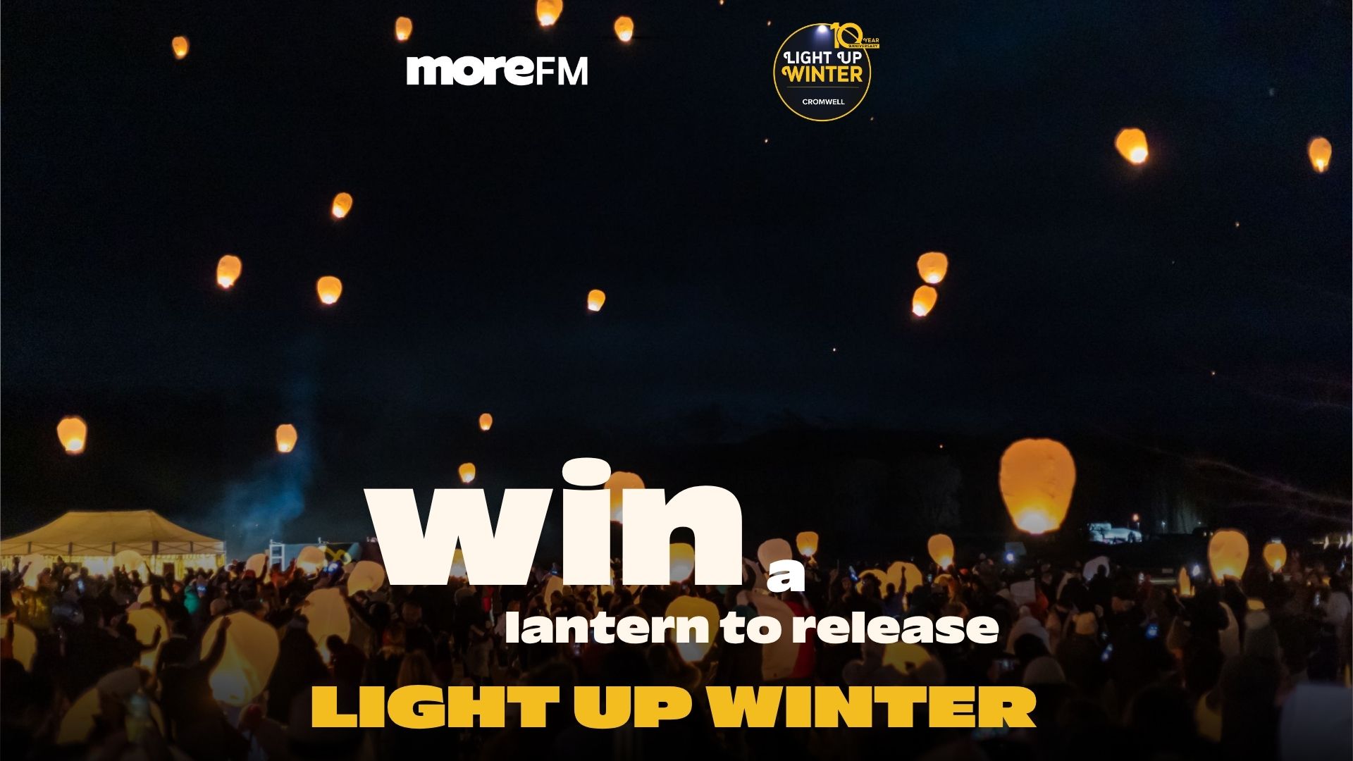 Numerous glowing orange sky lanterns ascend into a dark night sky above a crowd of people, with event branding for MoreFM and Light Up Winter Cromwell visible.