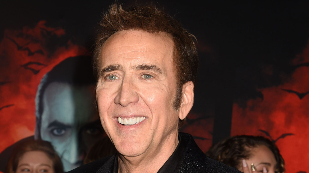 Actor Nicolas Cage, a white man with spiky brown hair and light eyes, smiles widely showing his teeth in front of a red, bat-filled movie poster with a blurry face.