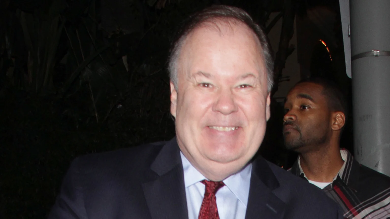 A smiling middle-aged white man in a suit and red tie looks forward, while a young Black man in a plaid shirt is partially visible behind him looking left, against a dark, blurry background.