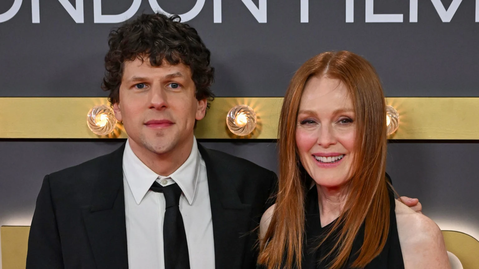 Jesse Eisenberg in a suit and Julianne Moore in a dark top stand together and smile, with the woman's hand on the man's arm, against a dark and gold background.