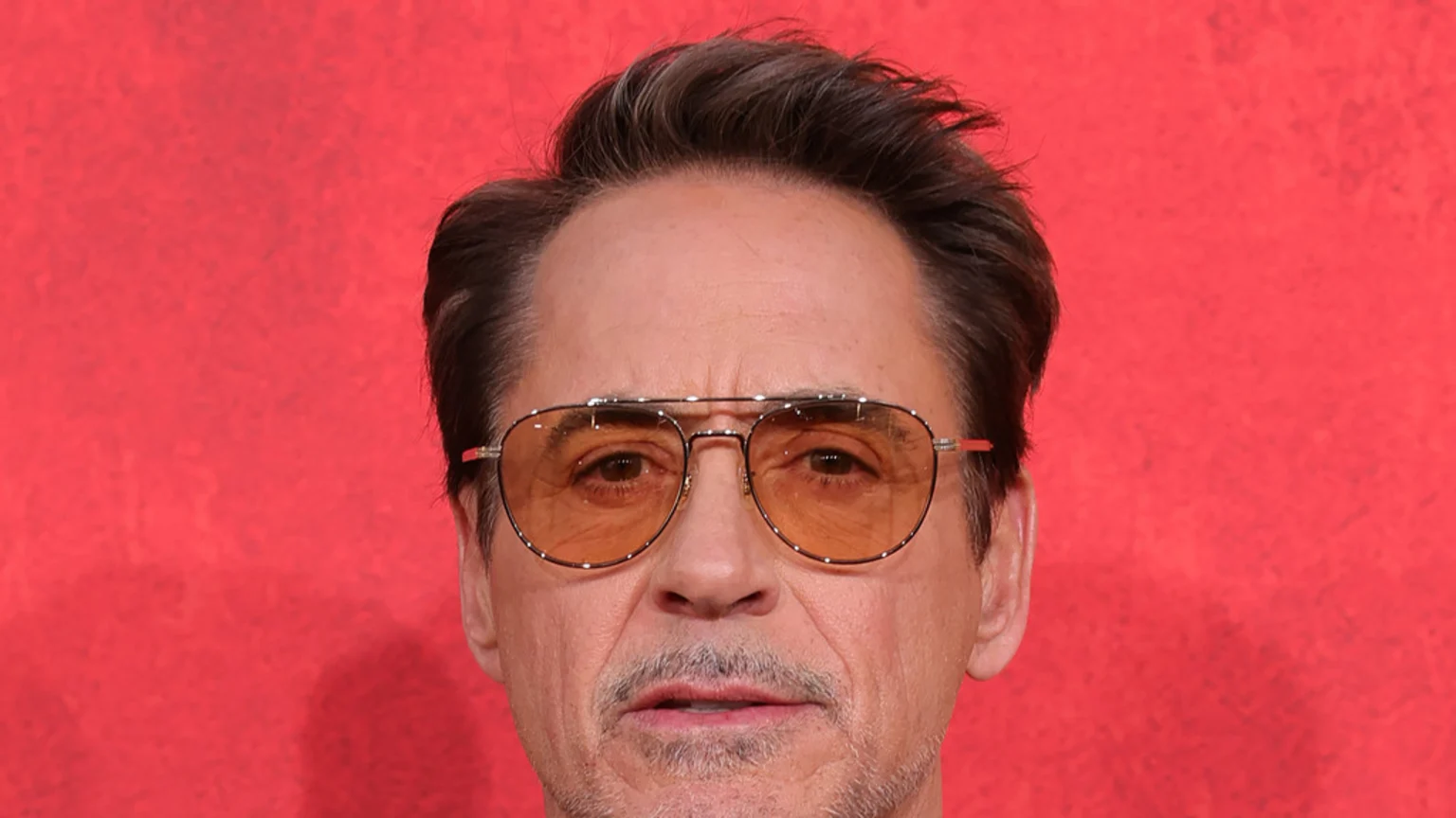Close-up of actor Robert Downey Jr. wearing orange-tinted aviator sunglasses and a goatee, with his hair styled upwards, against a red textured background.