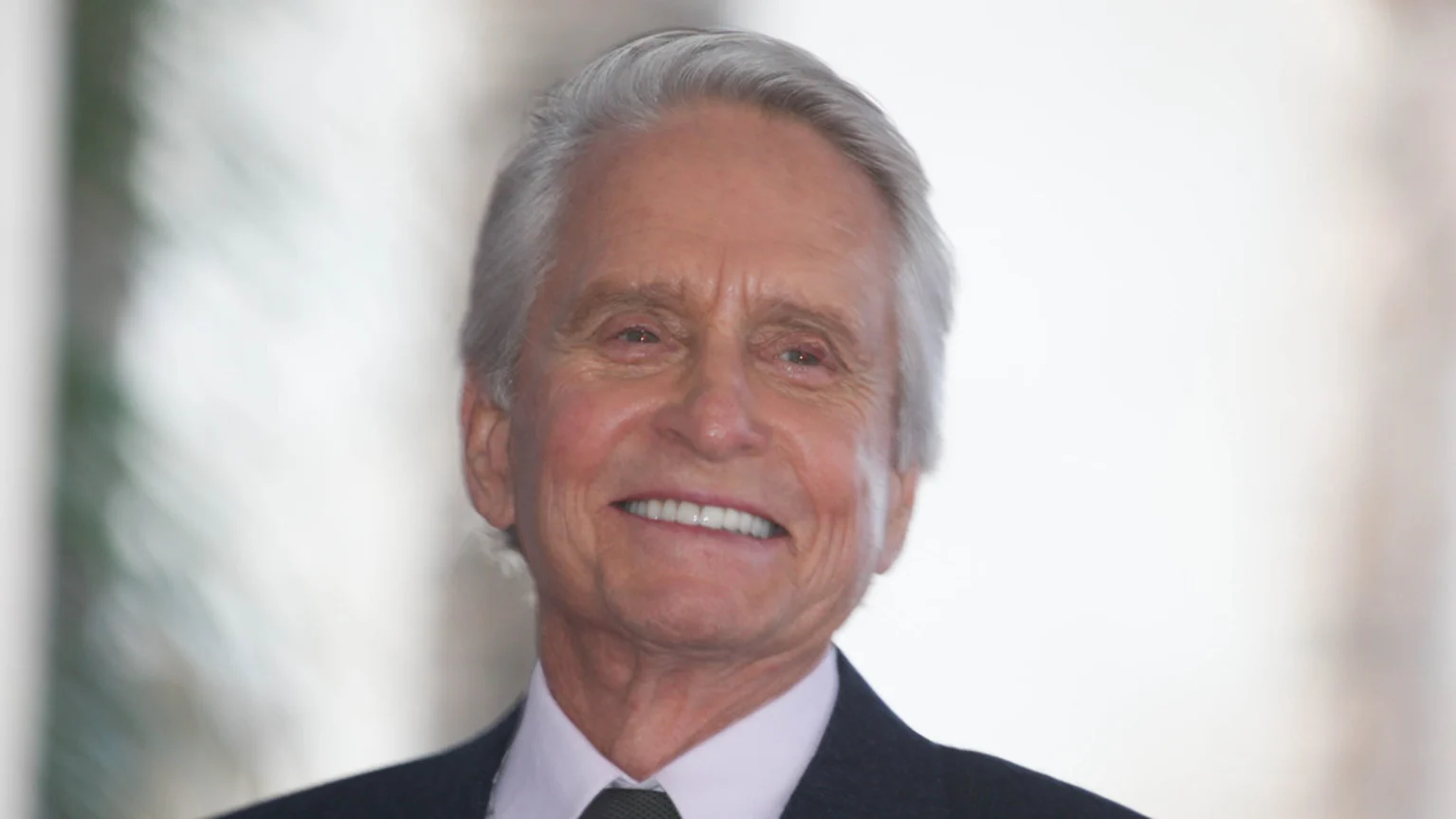 Actor Michael Douglas with gray hair smiles directly at the camera, wearing a dark suit and light shirt.