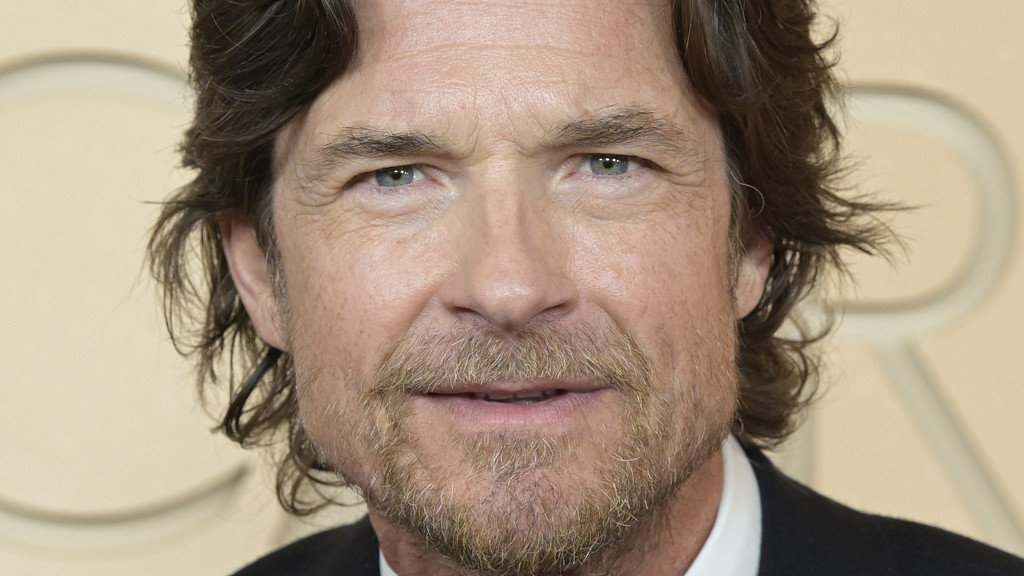 Close-up headshot of a smiling middle-aged man with wavy brown hair, light blue-grey eyes, and a light brown beard, wearing a dark suit and white shirt.