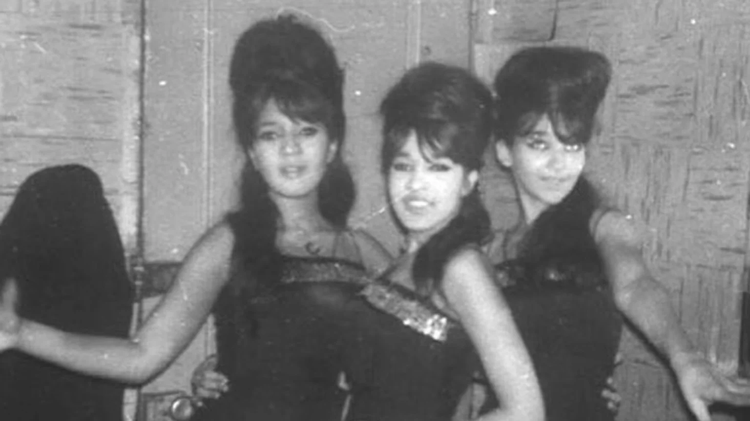 Black and white photo shows three women with prominent beehive hairstyles and dark, sparkling tops posing for the camera, with a dark figure partially visible on the left.