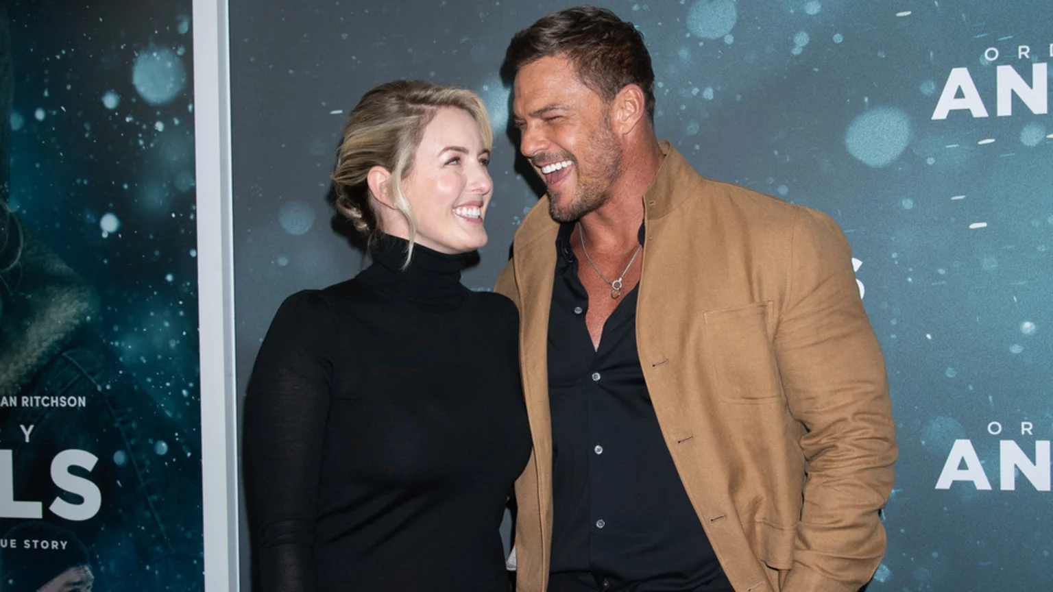 Alan Ritchson and Catherine Ritchson smiling at each other in front of movie posters with a dark teal, snowy background.