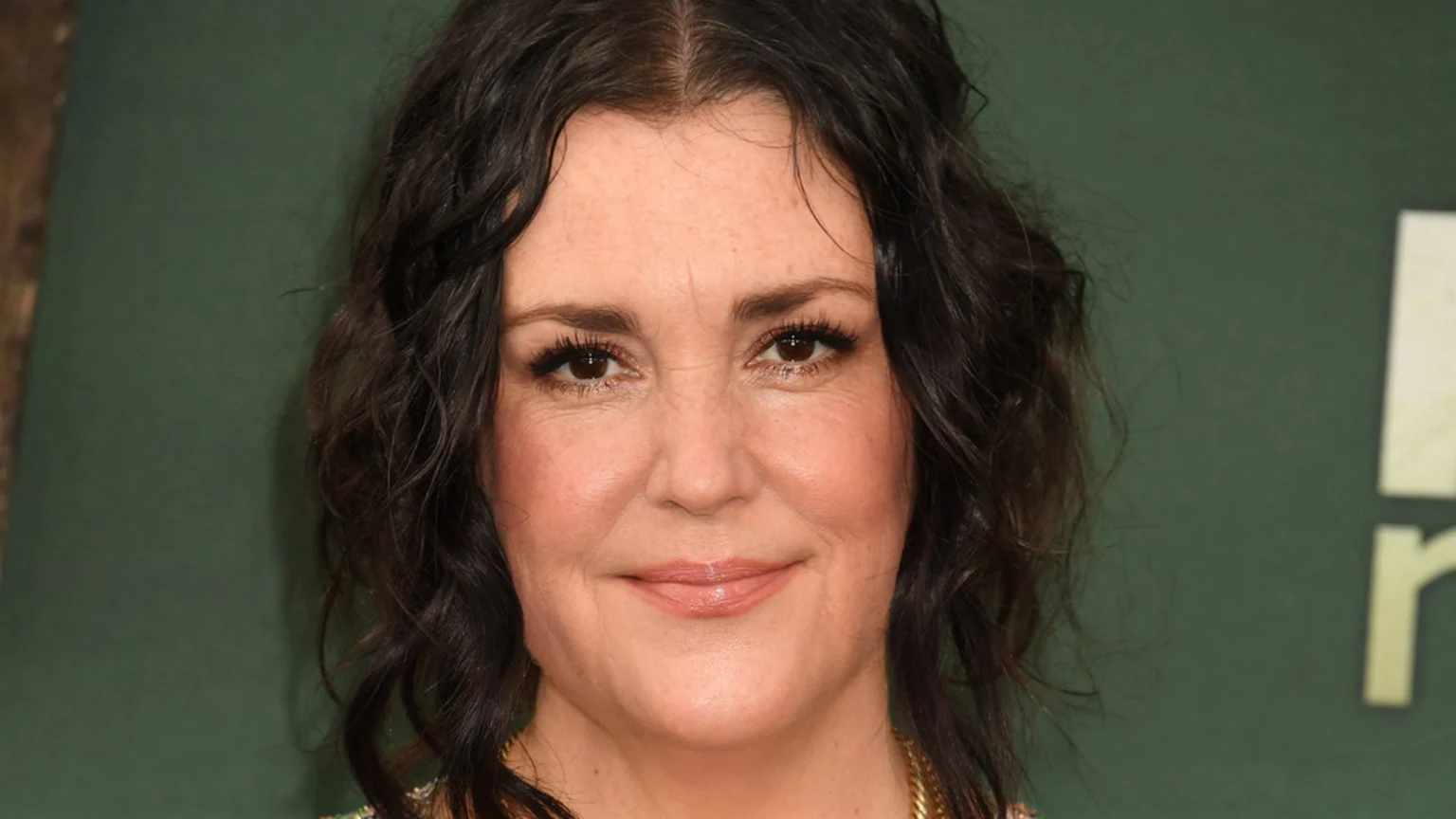 Close-up portrait of Melanie Lynskey smiling gently, with dark wavy hair, brown eyes, and light makeup, against a dark green background.