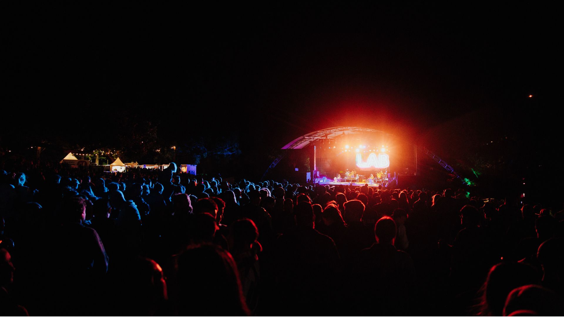A large crowd of people are gathered at night, watching a band perform on a brightly lit outdoor stage with "LAB" displayed, while tents and lights are visible in the distance.