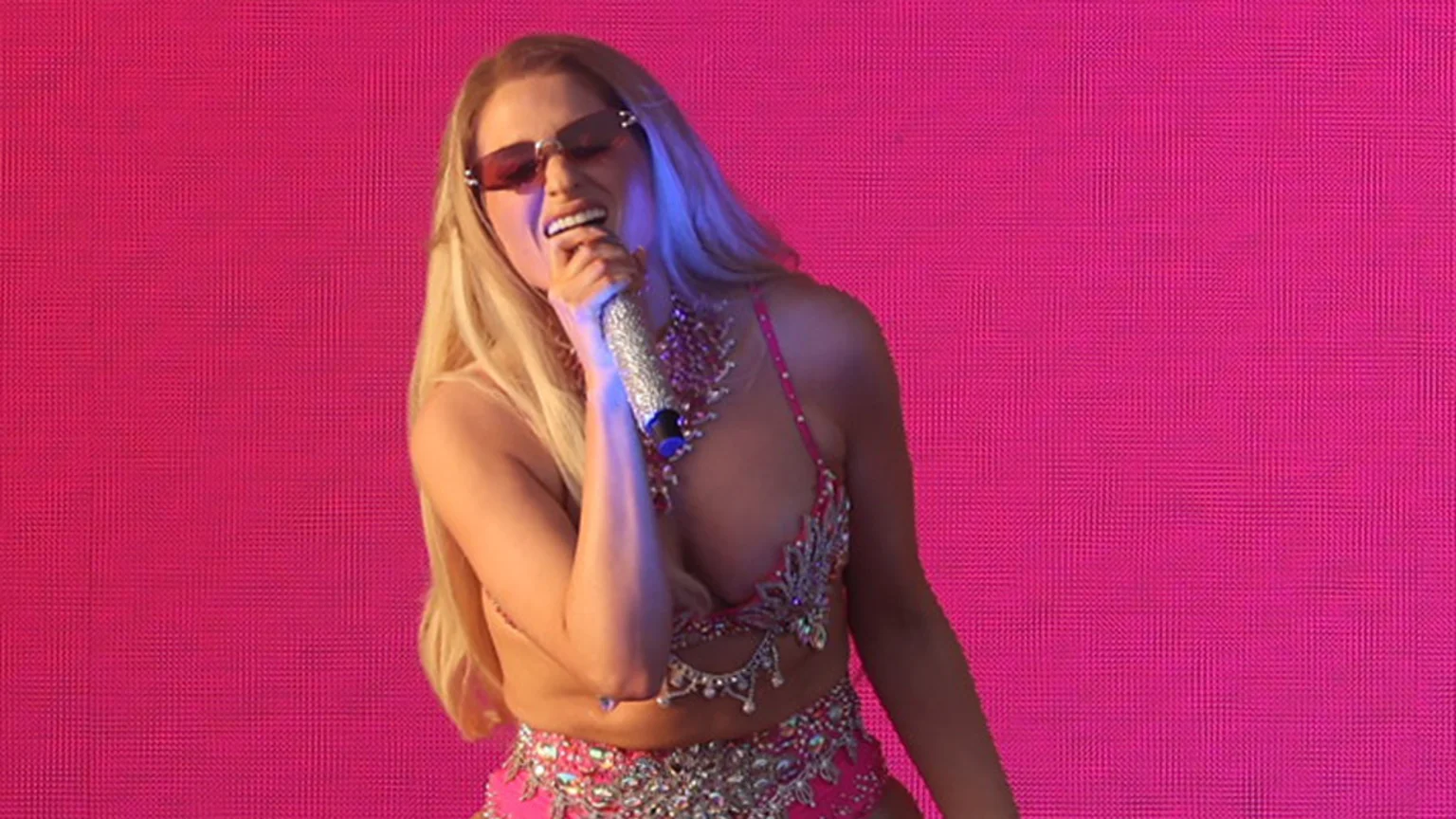A blonde woman in sunglasses sings into a sparkling microphone, wearing a pink, jewel-encrusted costume against a bright pink background, illuminated by a blue light.