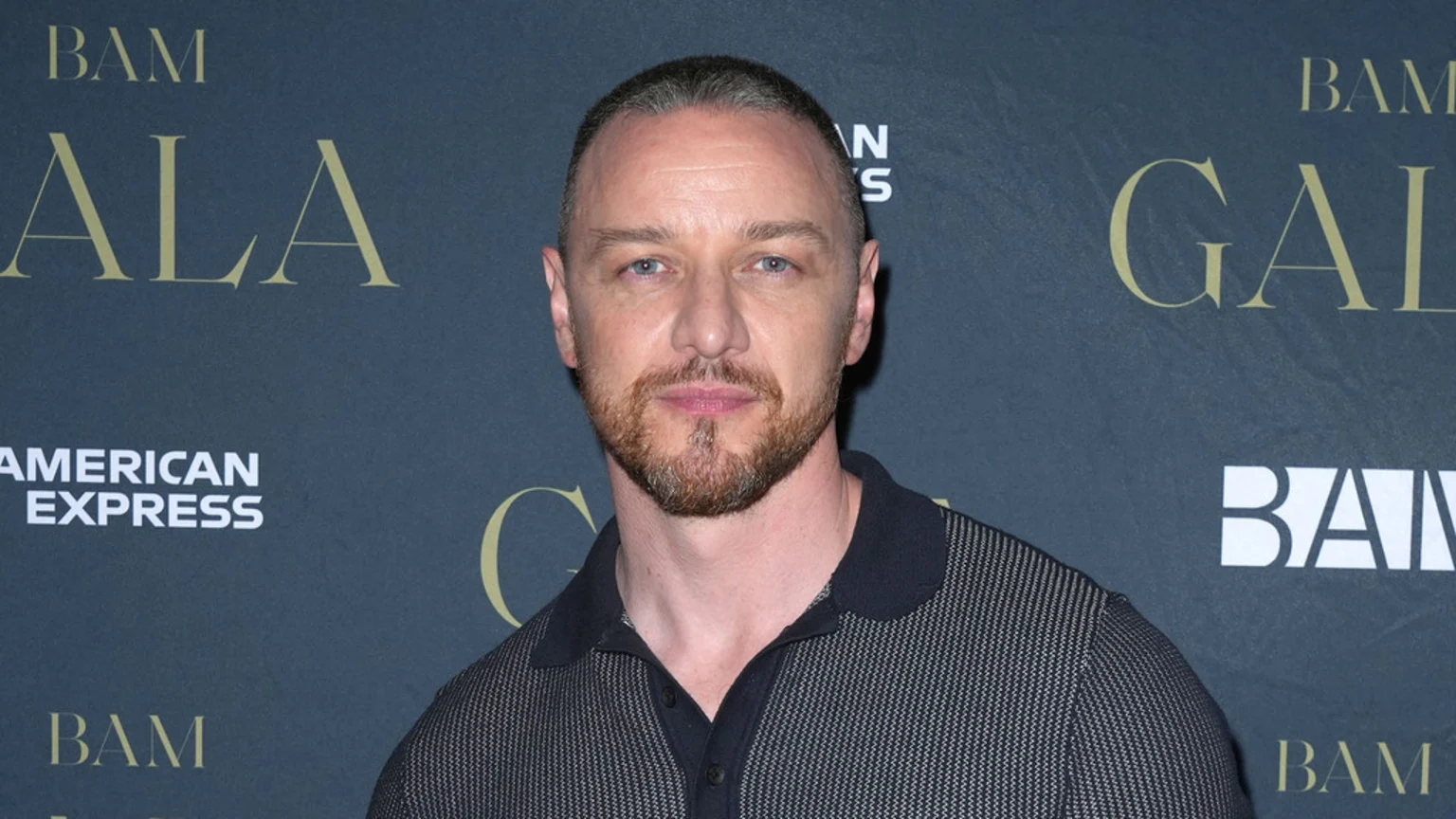 Actor James McAvoy with a short buzzcut and beard, wearing a dark textured polo shirt, poses against a dark backdrop featuring "BAM GALA" and "AMERICAN EXPRESS" logos.