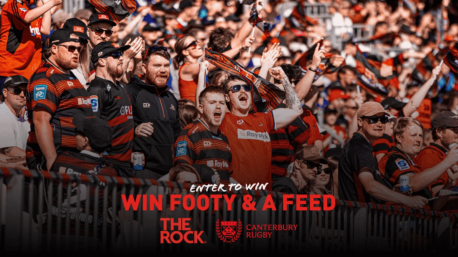 A large, enthusiastic crowd of rugby fans, many wearing red and black striped jerseys, cheer and wave flags in stadium stands.