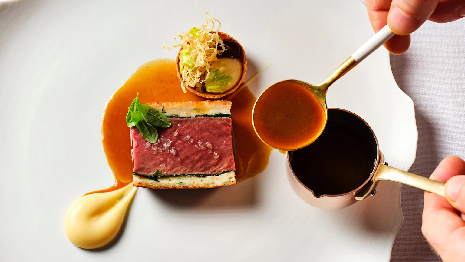 A gourmet dish on a white plate with a rectangular meat item topped with salt and leaves, a dollop of yellow puree, and a small tartlet with various fillings, as a hand pours a brown sauce from a copper pot.