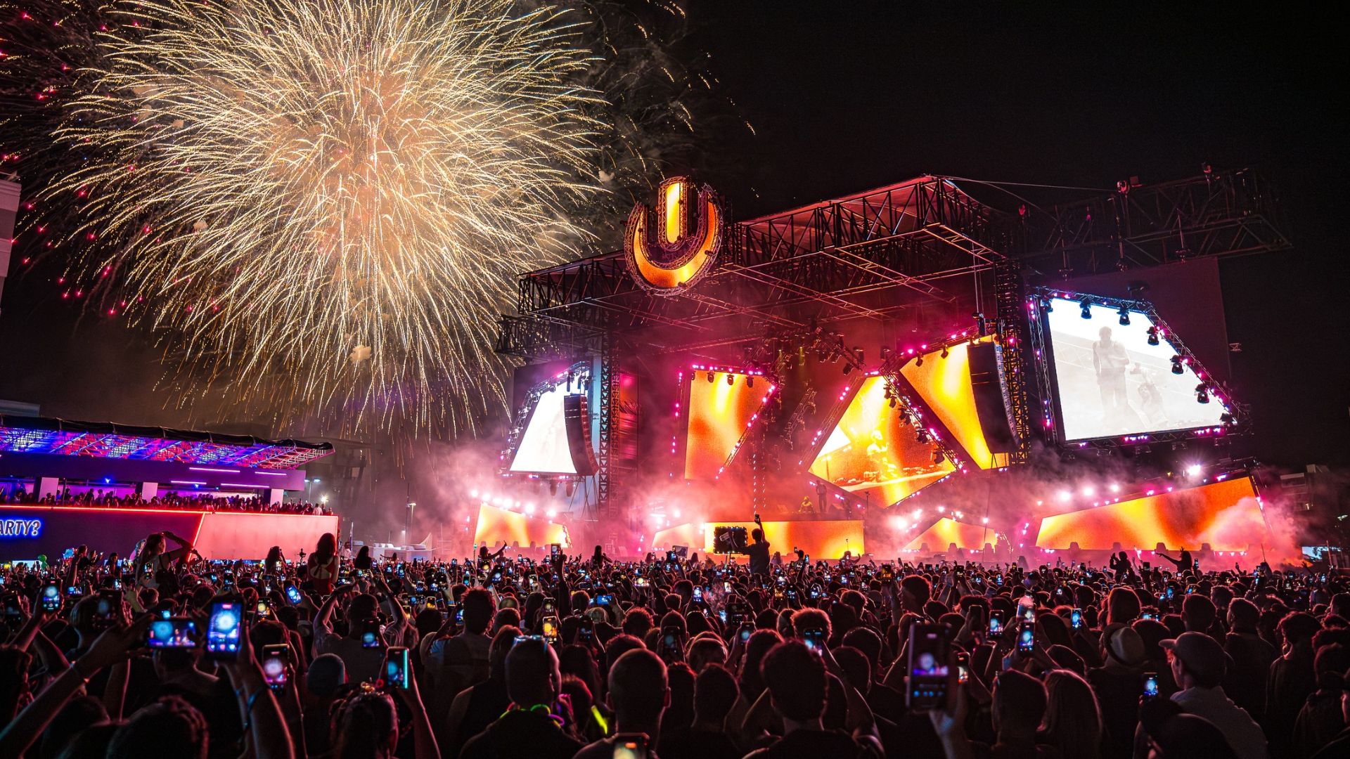 An outdoor night concert with a huge stage displaying the Ultra logo, vibrant lights, and large video screens, while a vast crowd holds up phones and fireworks burst overhead.