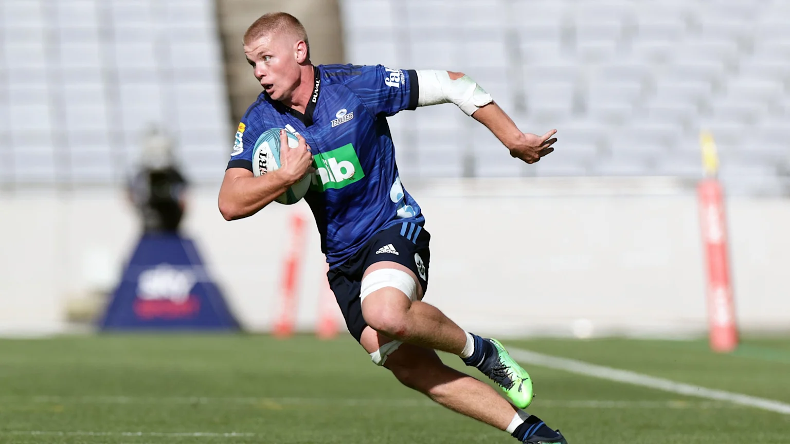 A male rugby player in a blue "Blues" jersey, black shorts, and bandages, runs on a green field while carrying a light blue rugby ball; stadium seating is visible in the background.