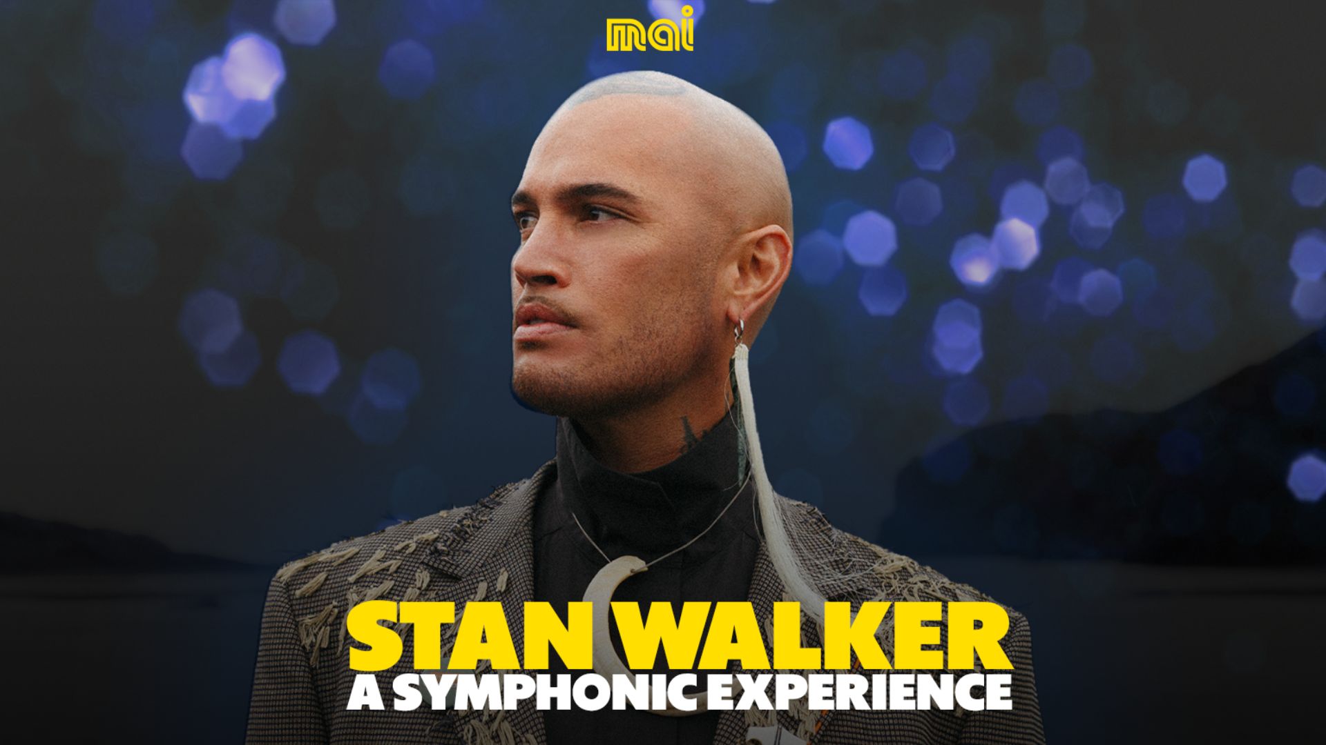 Stan Walker, a man with a shaved head and feather earring, looks left in a textured jacket, with "STAN WALKER A SYMPHONIC EXPERIENCE" in yellow text below him against a dark, bokeh background.
