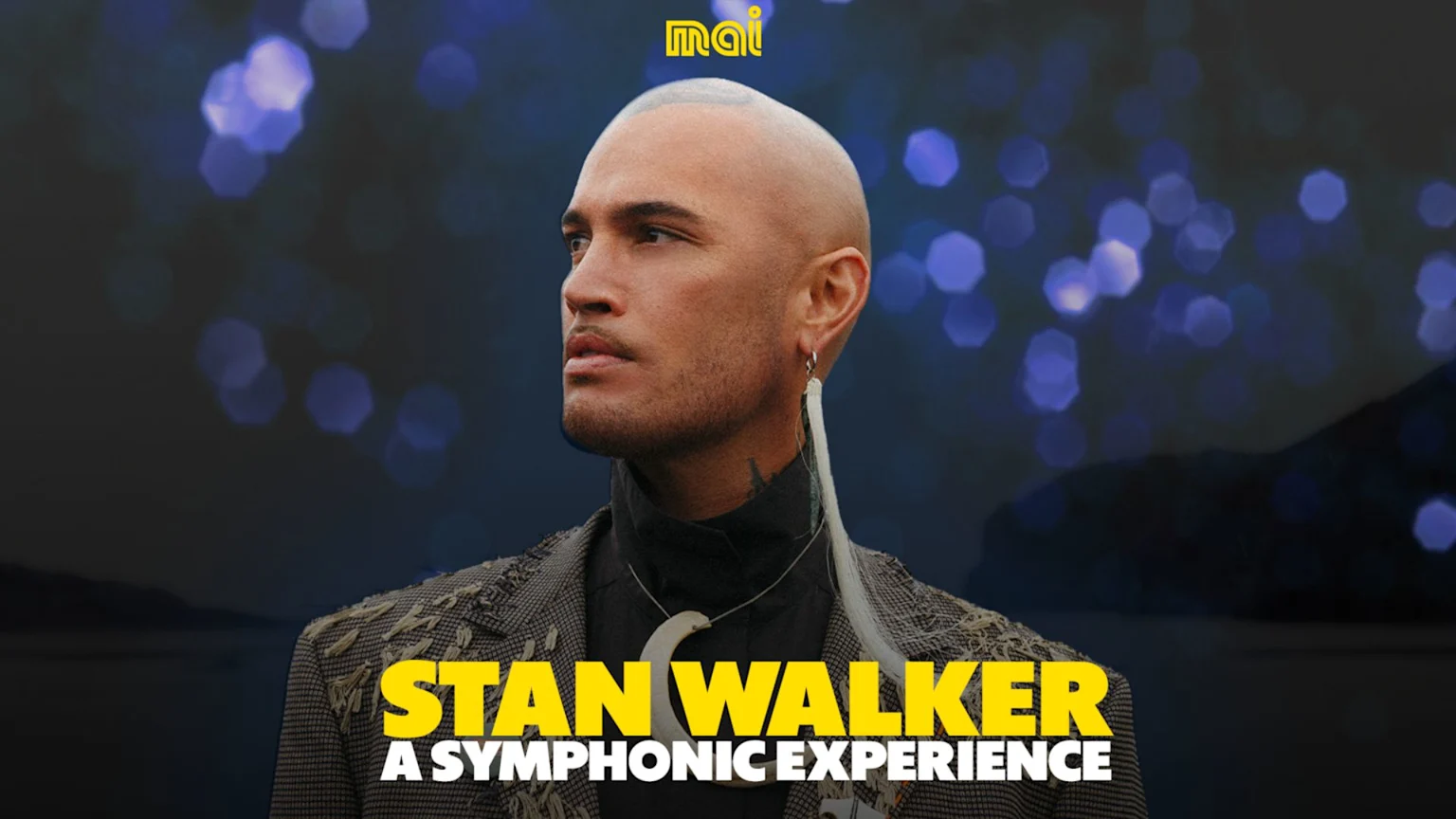Stan Walker, a man with a shaved head and feather earring, looks left in a textured jacket, with "STAN WALKER A SYMPHONIC EXPERIENCE" in yellow text below him against a dark, bokeh background.