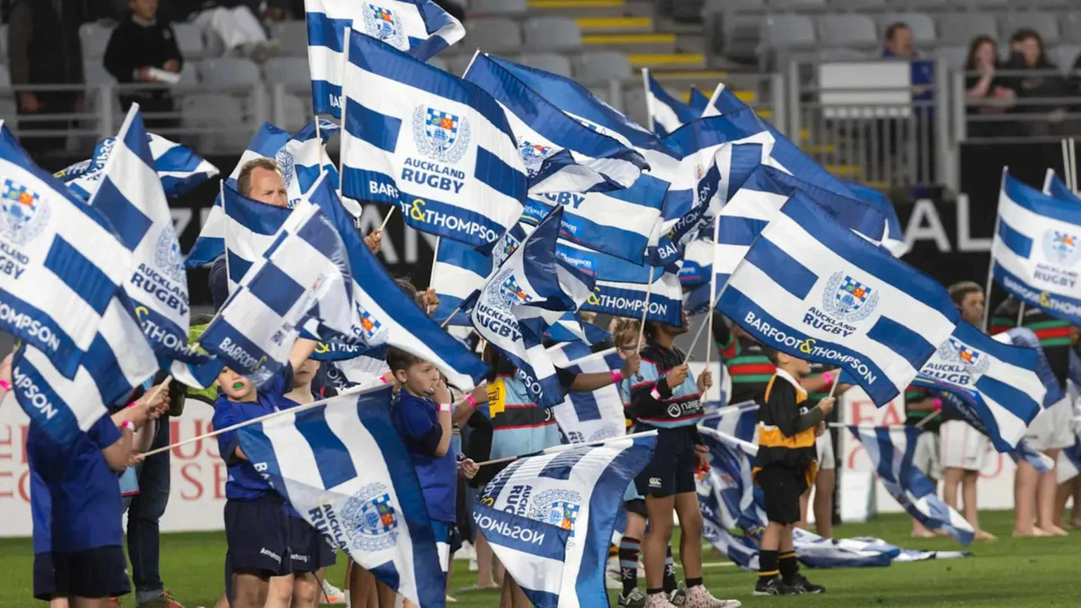 Children and adults on a sports field wave numerous blue and white Auckland Rugby flags, some with Barfoot & Thompson branding.