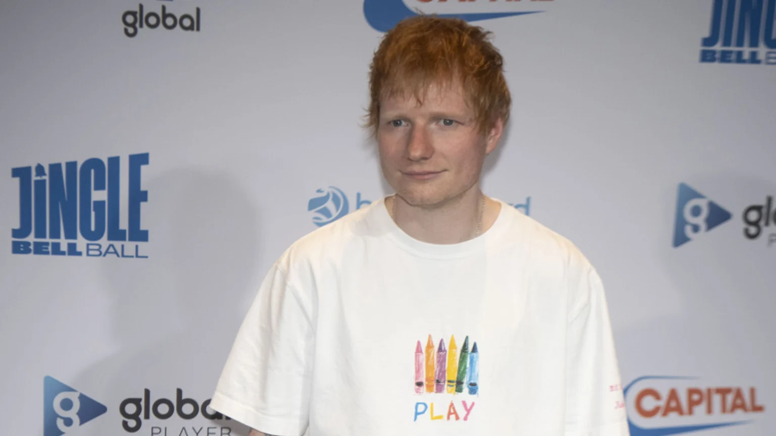 Ed Sheeran, with red hair and a subtle smile, wears a white t-shirt with a colorful crayon design and the word "PLAY" on it, standing against a white backdrop with event logos.