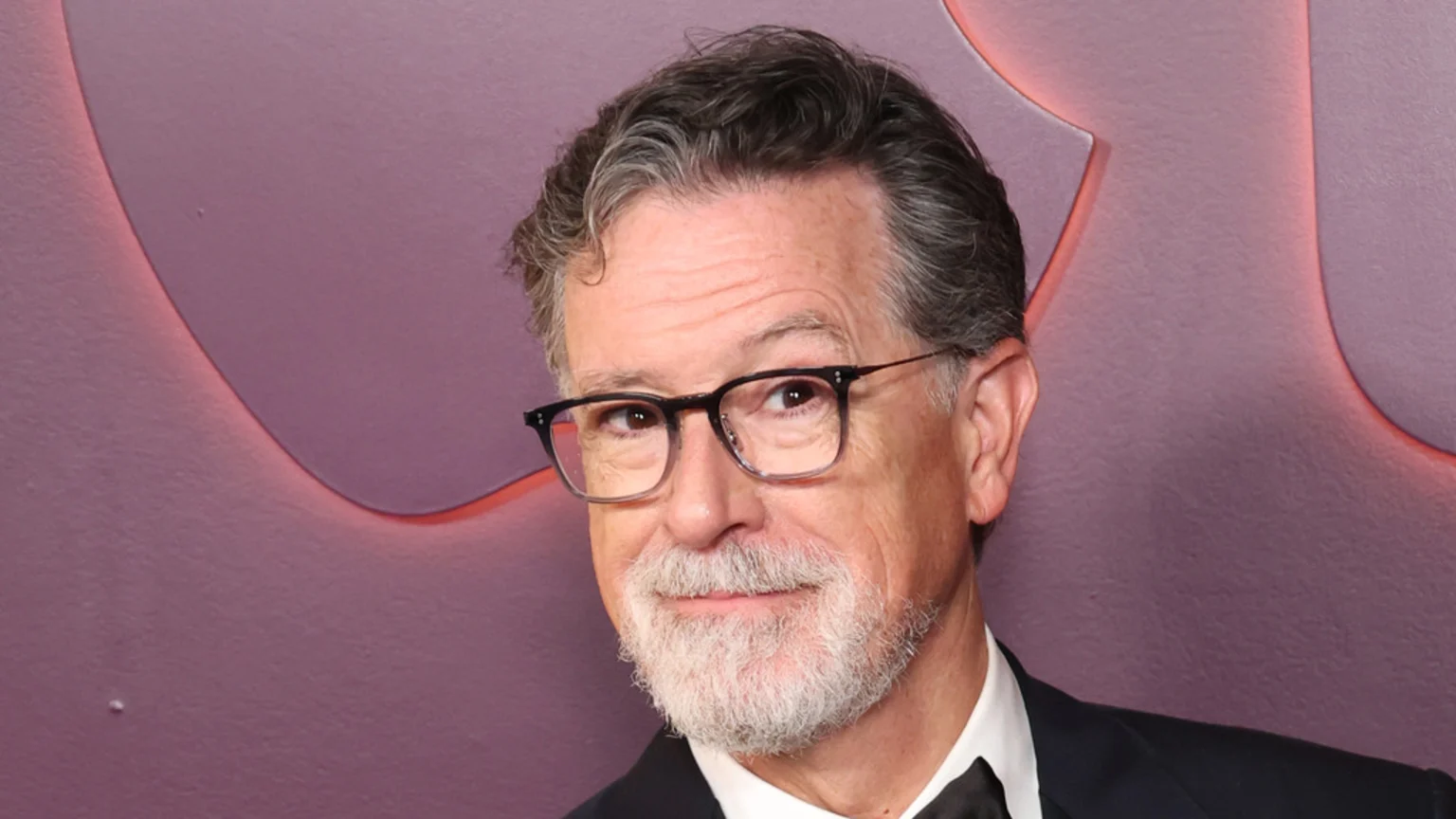 Stephen Colbert, a white man with glasses, grey hair, and a beard, smiles slightly while wearing a suit against a blurry purple background.
