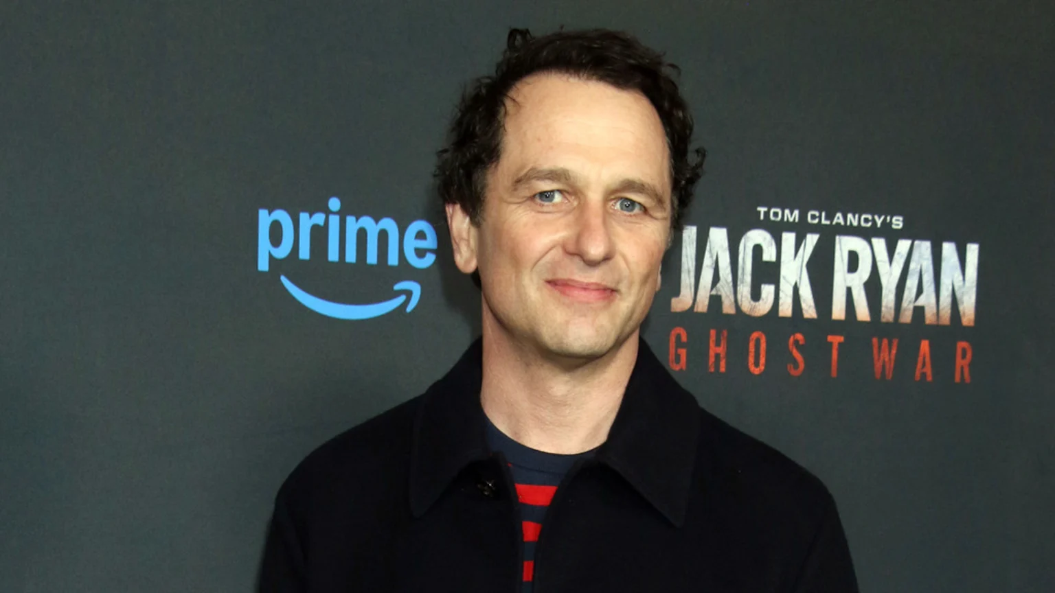 Matthew Rhys smiles, wearing a dark jacket over a striped shirt, in front of a backdrop featuring the Amazon Prime logo and "Tom Clancy's Jack Ryan Ghost War."