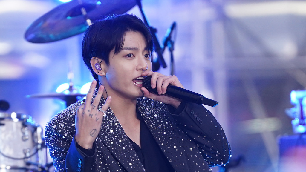 A male singer with dark hair holds a microphone, showing "ARMY" finger tattoos, a lip piercing, and a purple in-ear monitor, wearing a sparkly blazer against a blurred blue background with drums.