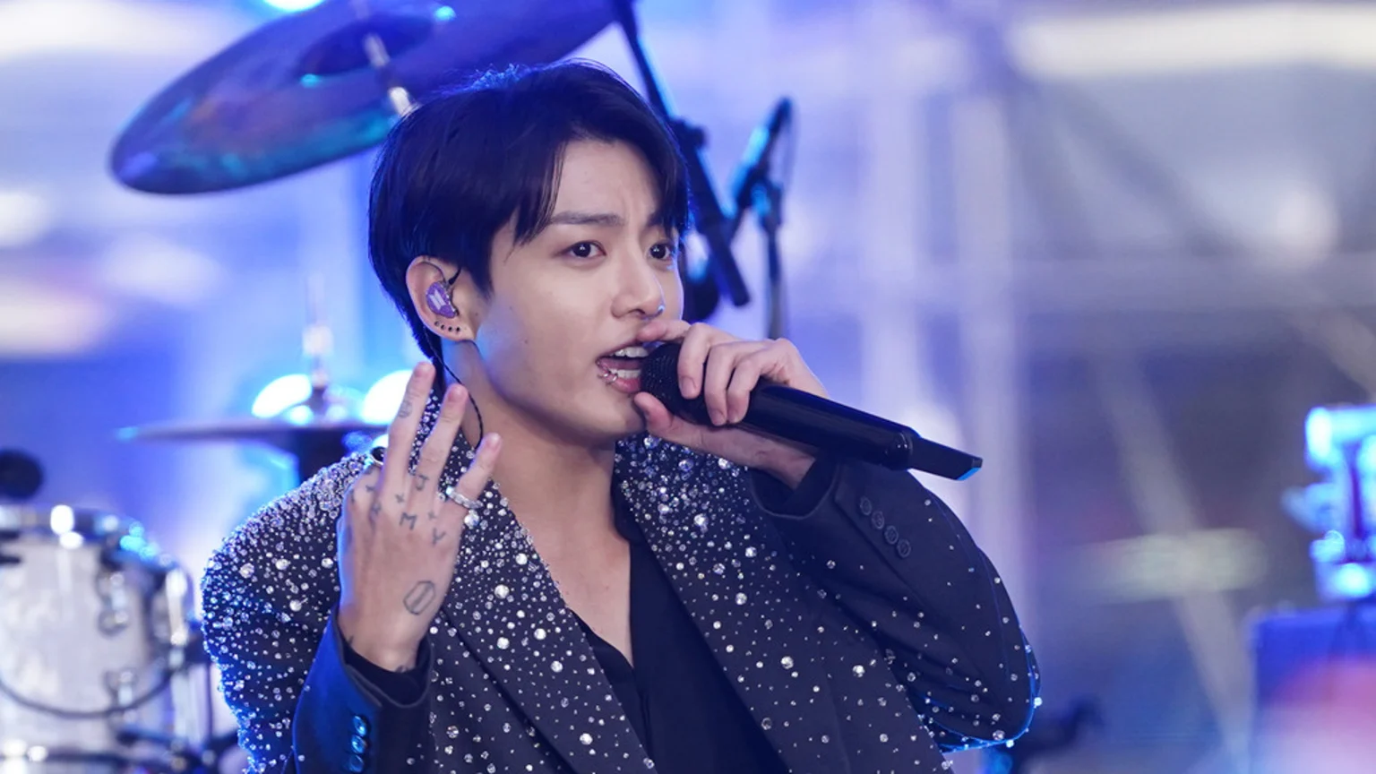 A male singer with dark hair holds a microphone, showing "ARMY" finger tattoos, a lip piercing, and a purple in-ear monitor, wearing a sparkly blazer against a blurred blue background with drums.