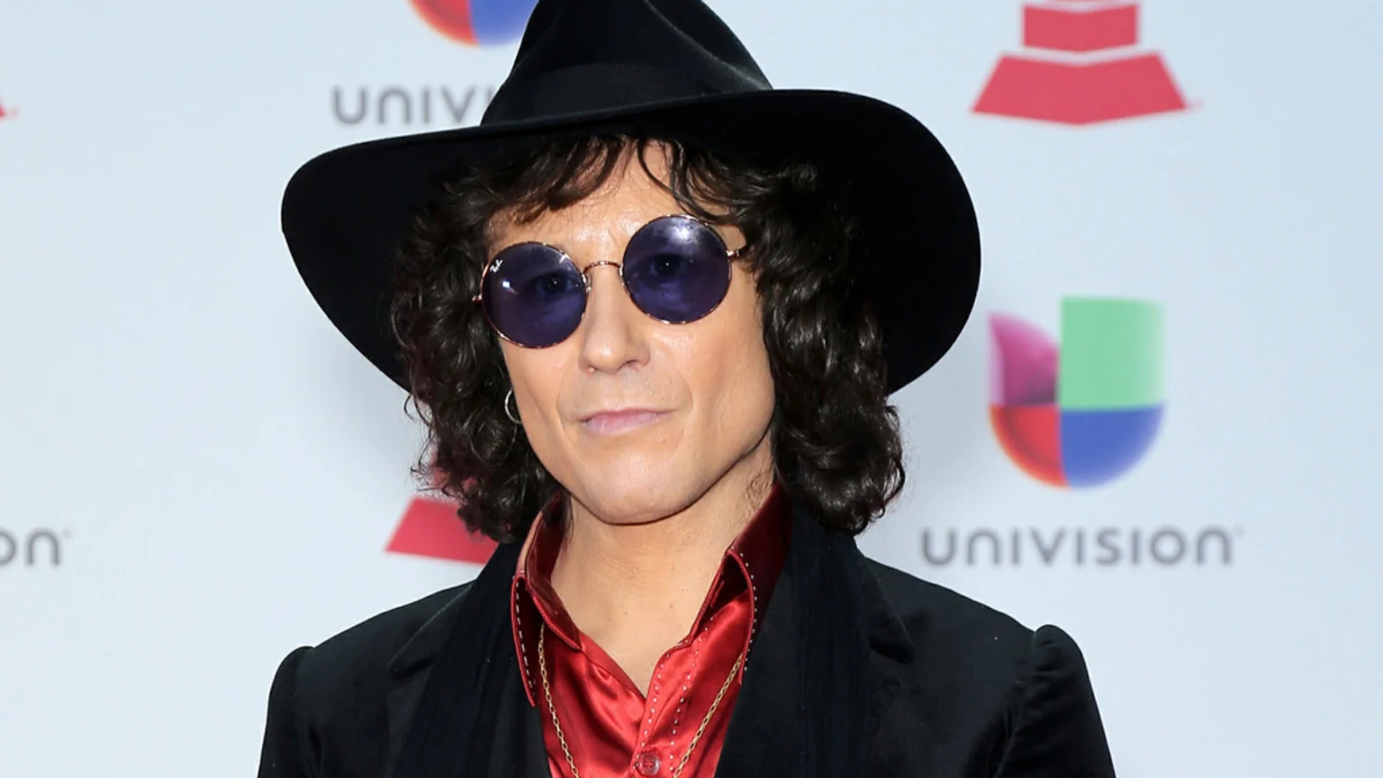 A man with dark curly hair wears a black hat, round purple sunglasses, a hoop earring, a gold chain, a red shirt, and a black jacket, against a white background with blurred logos including "UNIVISION".