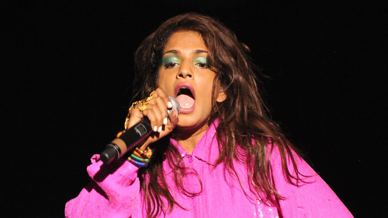 M.I.A. performs on stage, wearing a pink top, with bright green eyeshadow and her mouth open, holding a microphone.