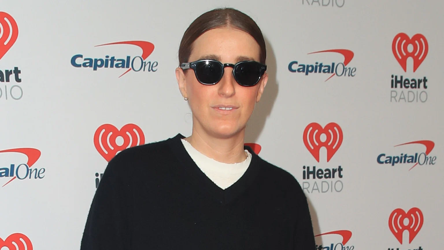 A person with light brown hair pulled back, wearing sunglasses and a black V-neck sweater over a white shirt, stands in front of a white wall adorned with Capital One and iHeart Radio logos.