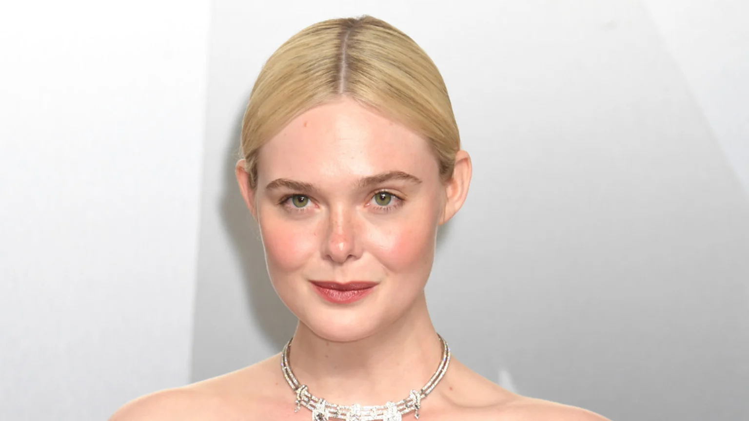 A blonde woman with green eyes and light skin, wearing a sparkling silver necklace, smiles gently against a light background.
