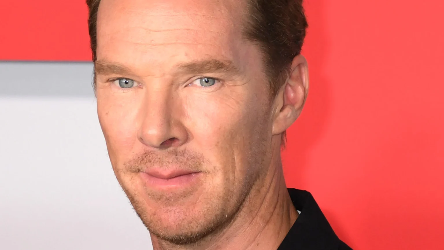 A close-up portrait of actor Benedict Cumberbatch, featuring his light blue eyes, light brown hair, and stubble, as he looks at the viewer with a subtle smile against a red and white background.