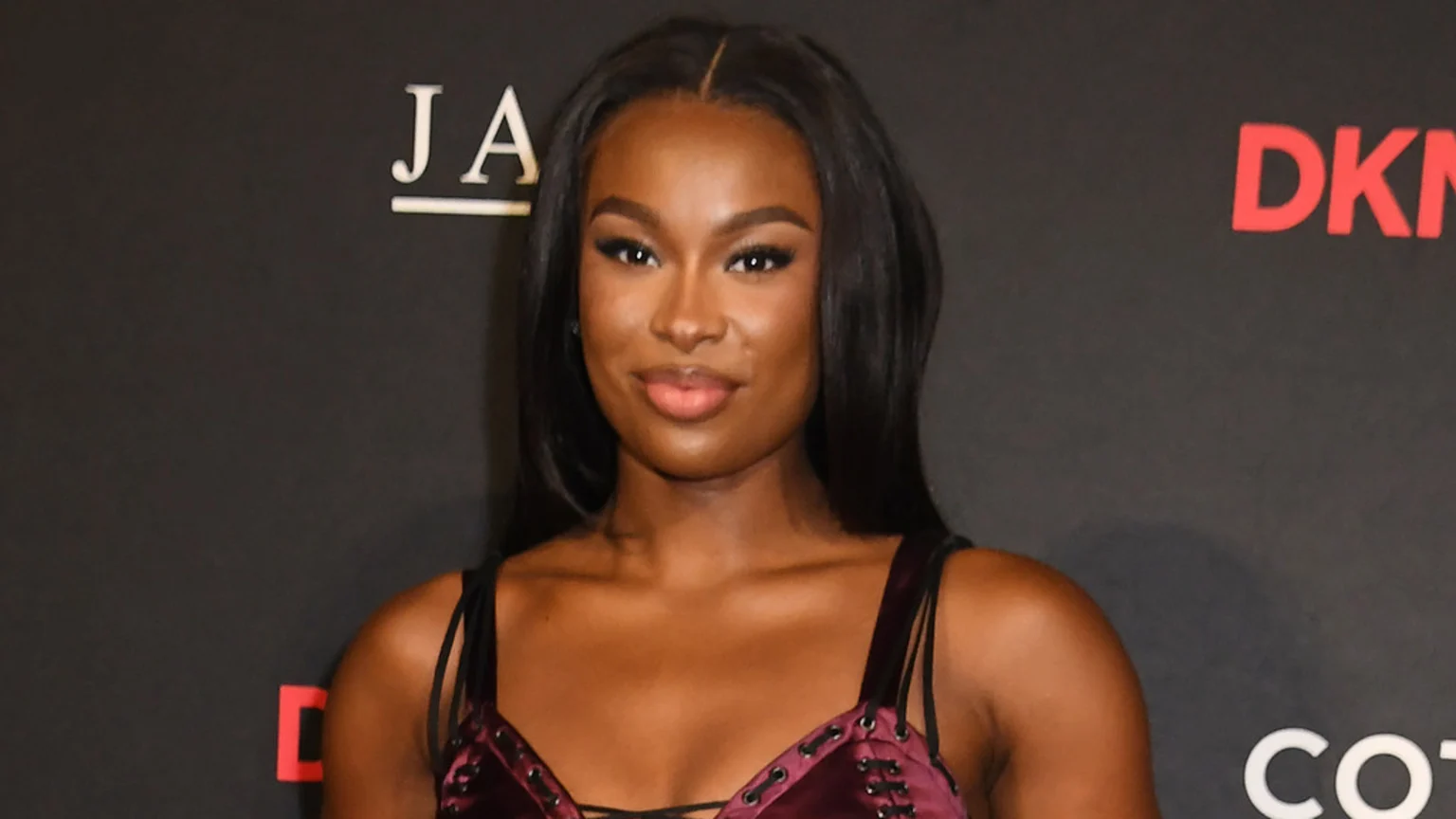 A close-up studio portrait features a smiling woman with dark skin and long, straight dark hair, wearing a dark red top with laced straps. The dark backdrop shows logos 'JA', 'DKN', and 'CO'.