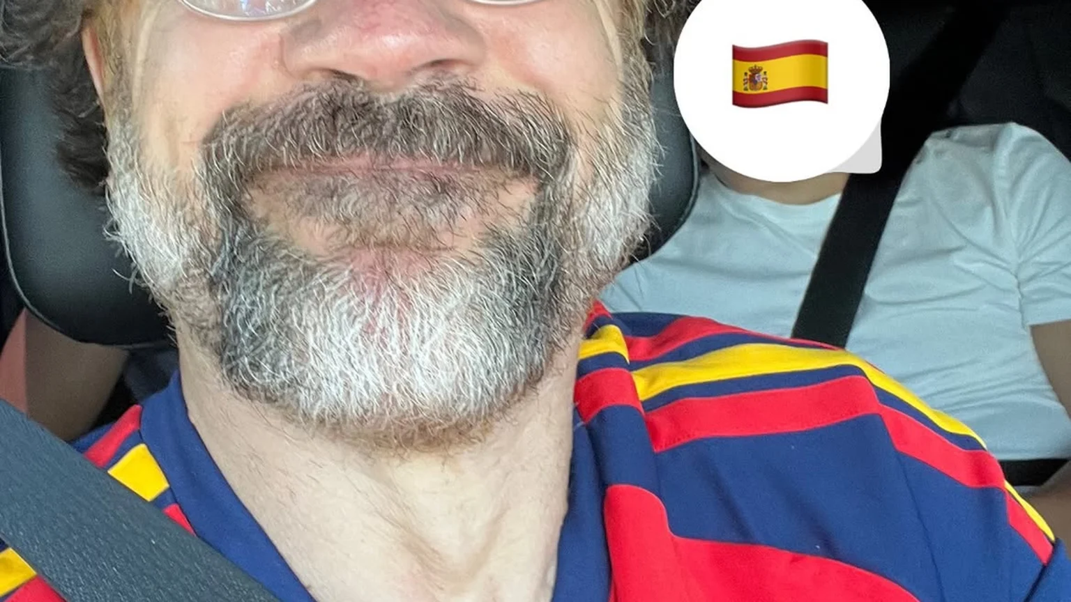 Javier Bardem, wearing a red and blue Spanish national team jersey, smiles at the camera in a car, with a Spanish flag emoji partly obscuring a person in the background.