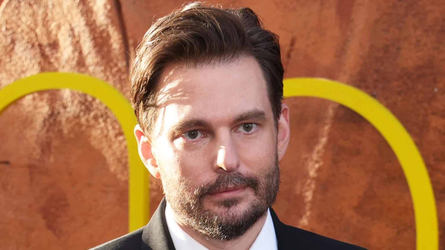 Sam Levinson with dark hair and a beard looks directly at the camera, wearing a suit jacket and white shirt, against a reddish-brown textured background with yellow arcs.