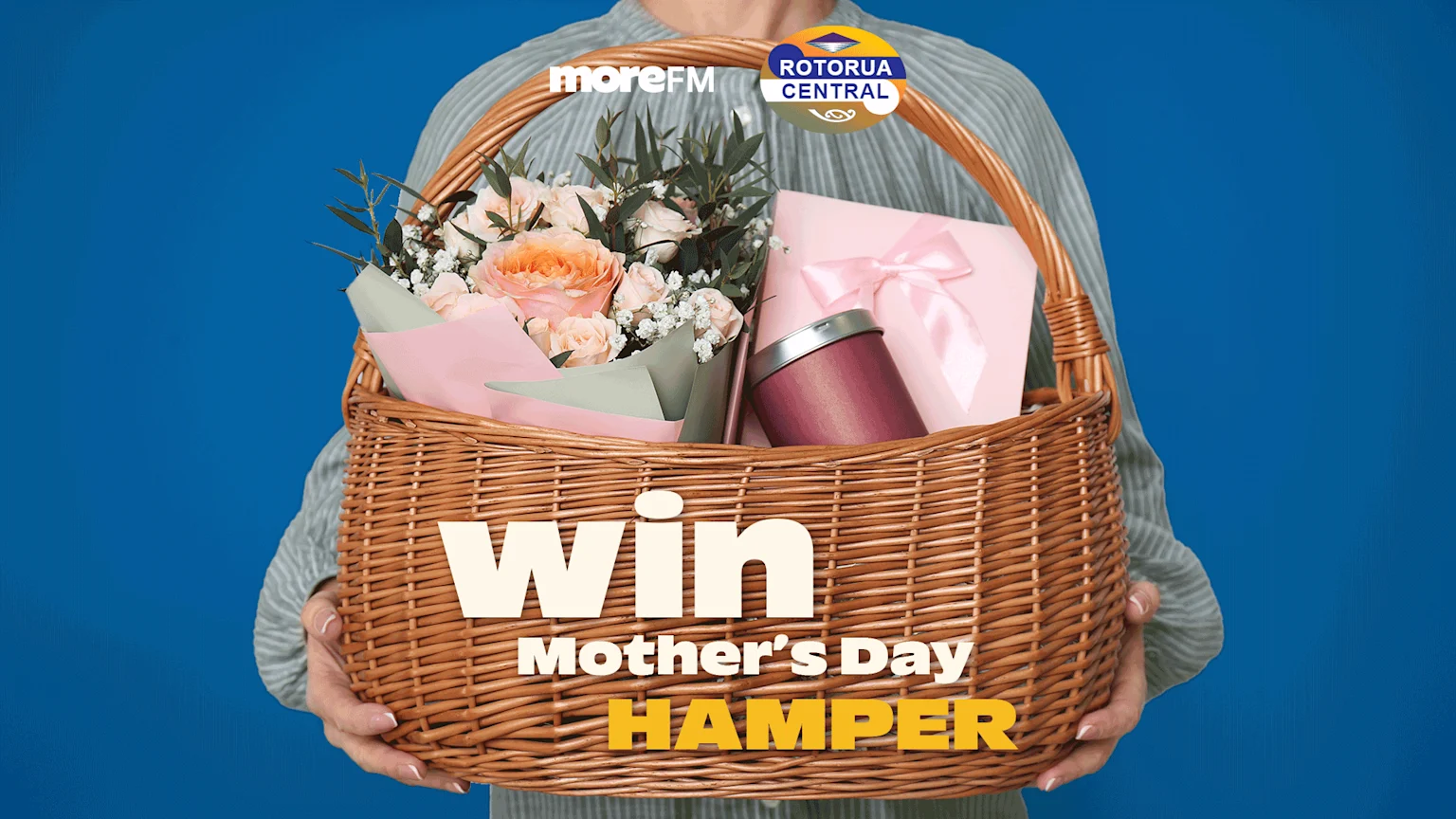 A person holds a wicker Mother's Day hamper filled with flowers, a gift box, and a canister, with "Win Mother's Day Hamper" text and "moreFM" and "Rotorua Central" logos.