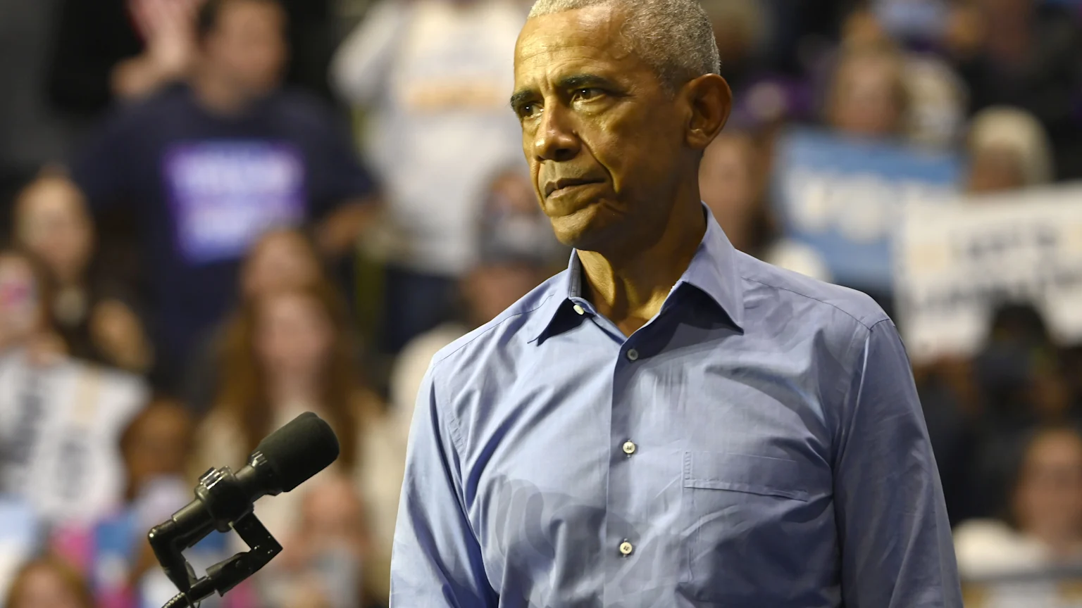 Barack Obama, with grey hair and a light blue shirt, looks to his left while standing in front of a blurred crowd, with a microphone in the foreground.