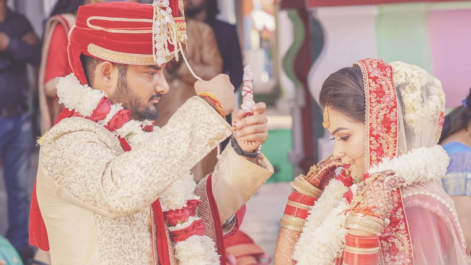 An Indian groom in a gold sherwani and red turban holds a ceremonial object, while the bride, adorned in a red and gold lehenga with a white floral veil and bangles, looks down during their wedding.