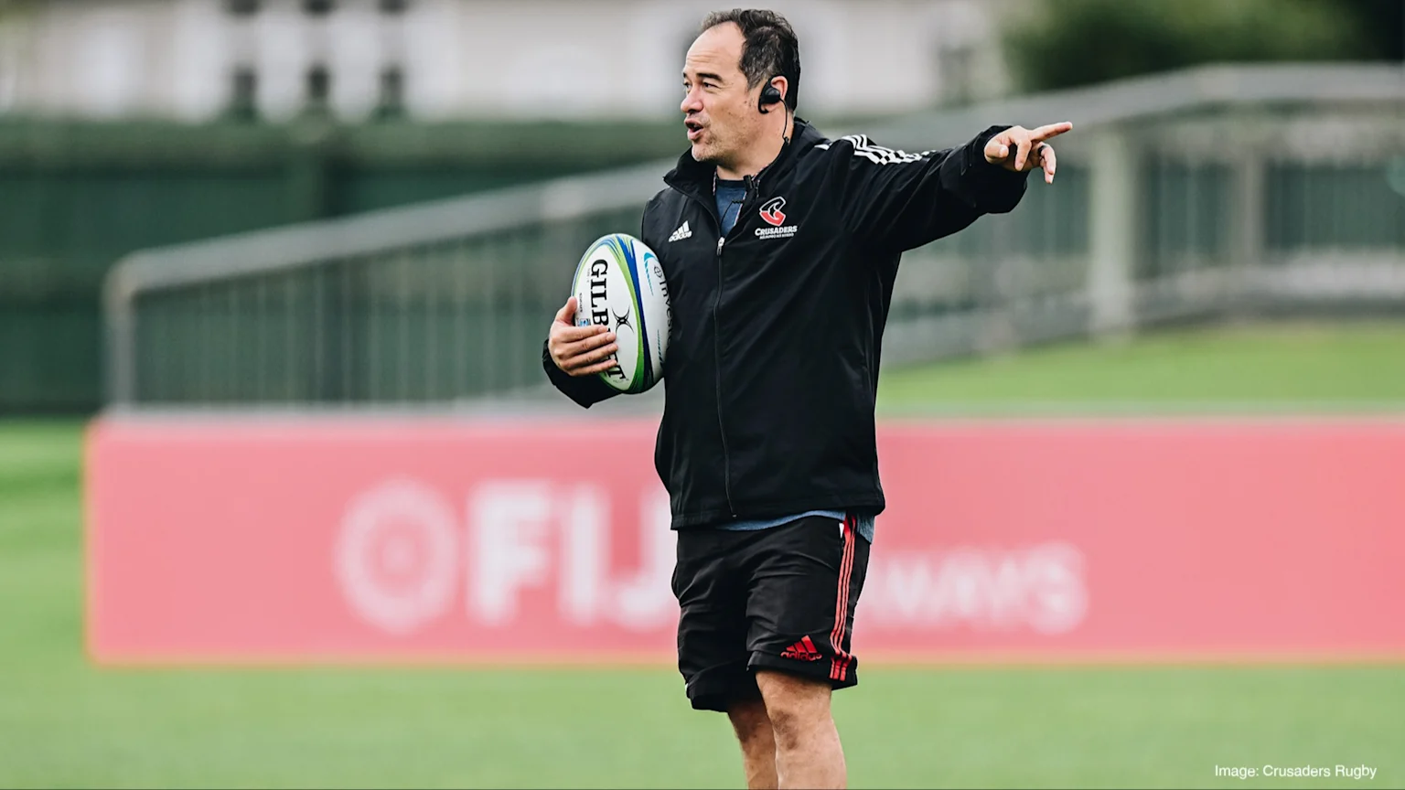 Scott Hansen, wearing a black Crusaders jacket, holds a rugby ball and points to the right on a green field with a red banner.