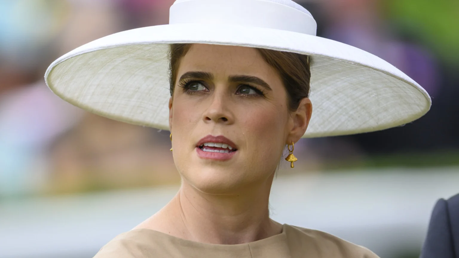 A close-up shows Princess Eugenie wearing a white wide-brimmed hat, a beige top, and gold mushroom earrings, looking upwards with her mouth slightly open.