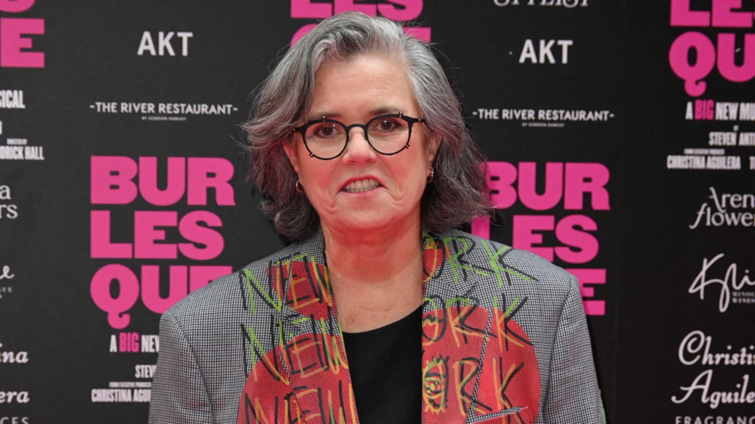 Rosie O'Donnell, with gray hair and glasses, smiles at the camera, wearing a gray checkered blazer with colorful "NEW YORK" graffiti, against a dark banner with "BURLESQUE" in pink.
