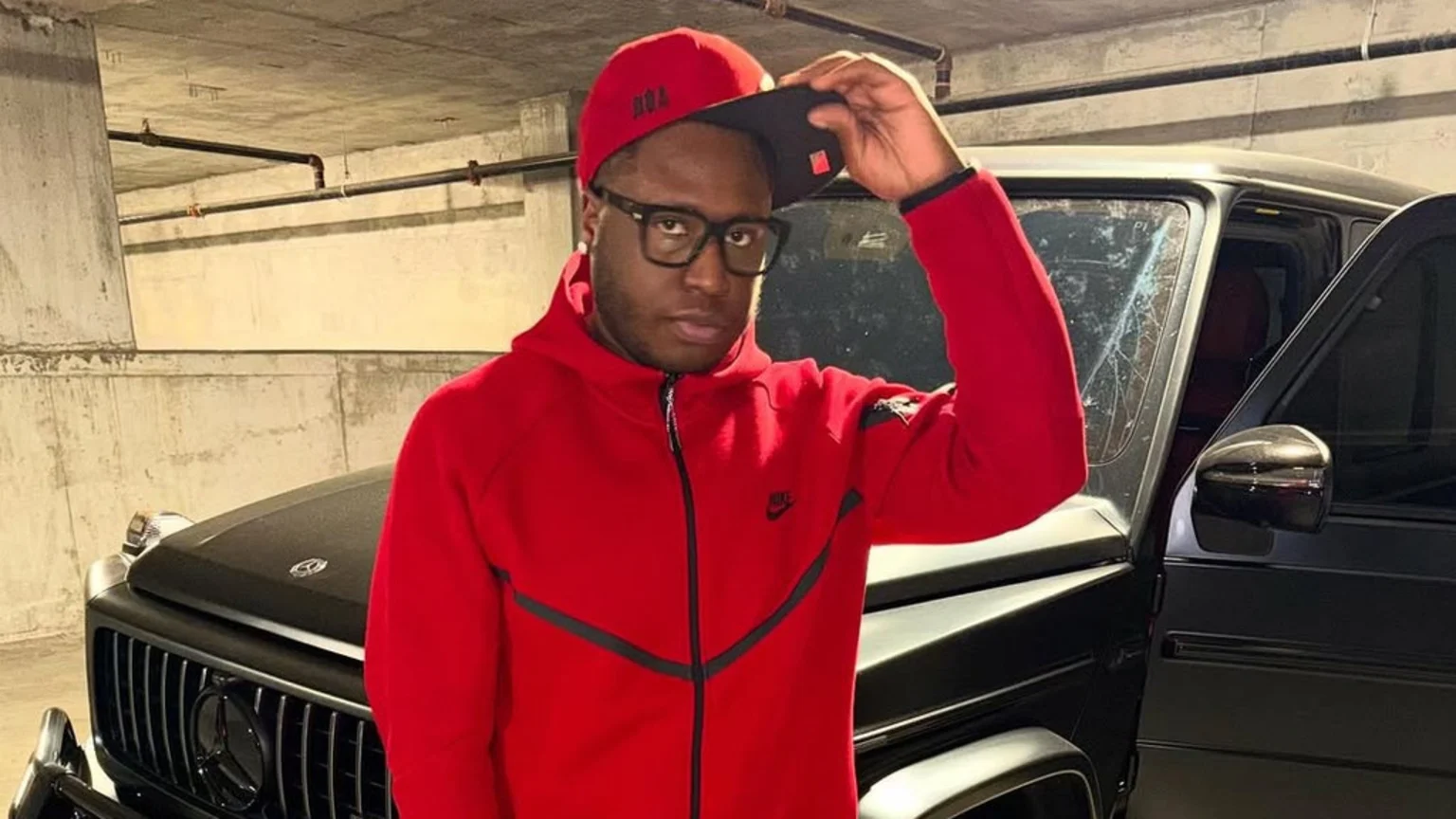 A man in a red hoodie, glasses, and a red cap poses in front of a black Mercedes-Benz G-Wagen with a cracked windshield in a parking garage.