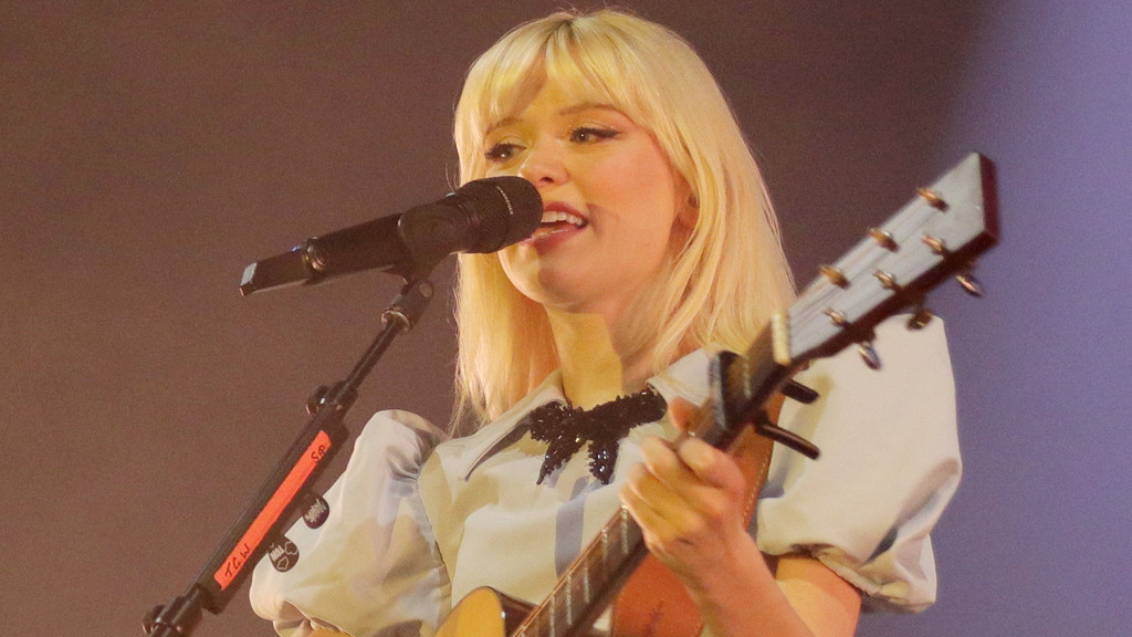 Maisie Peters, a young woman with blonde hair, sings into a microphone while holding an acoustic guitar on a softly lit stage.