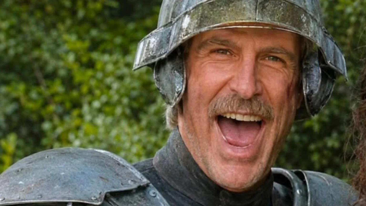 A man with a mustache, wearing a distressed metal helmet and shoulder armor, smiles widely with his mouth open, against a blurred green background.