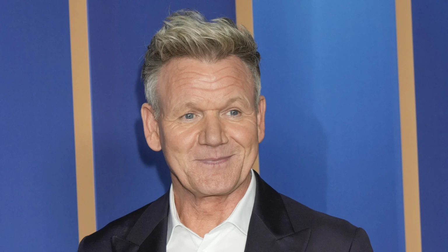 A smiling Gordon Ramsay with grey hair and a dark suit stands against a blue background with a vertical tan stripe.