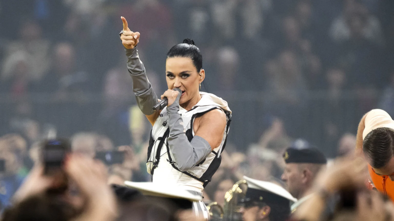 Katy Perry, with her hair in a bun, sings into a microphone while pointing her right index finger upwards, wearing a white and black vest and gray arm sleeves, with a blurred audience in the background.