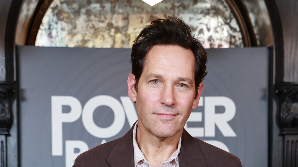 Paul Rudd, a man with dark hair and green eyes, looks directly at the camera with a faint smile, wearing a brown jacket over a striped shirt, against a grey banner with white text.