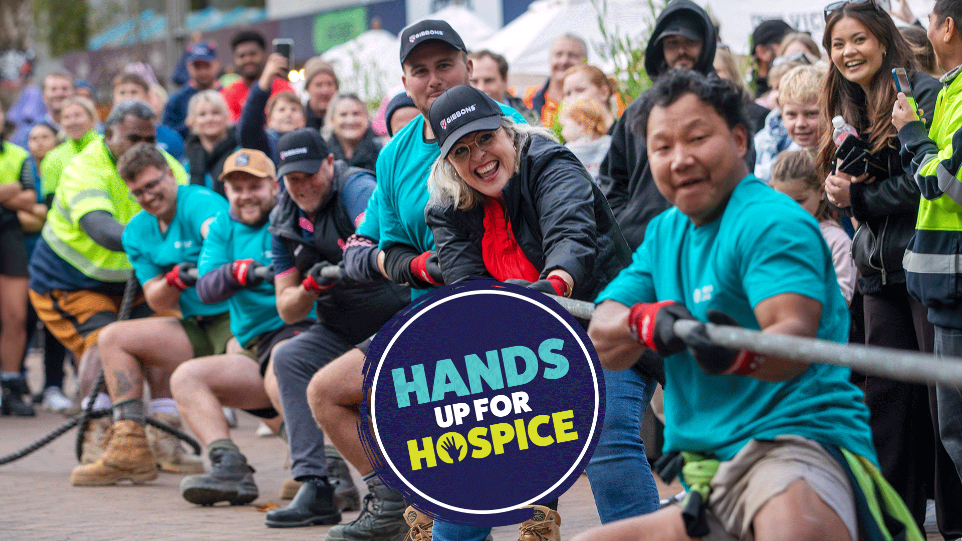 A diverse group of enthusiastic adults participates in a tug-of-war, pulling a thick rope with smiles, while a circular "Hands Up For Hospice" logo is overlaid in the center foreground.