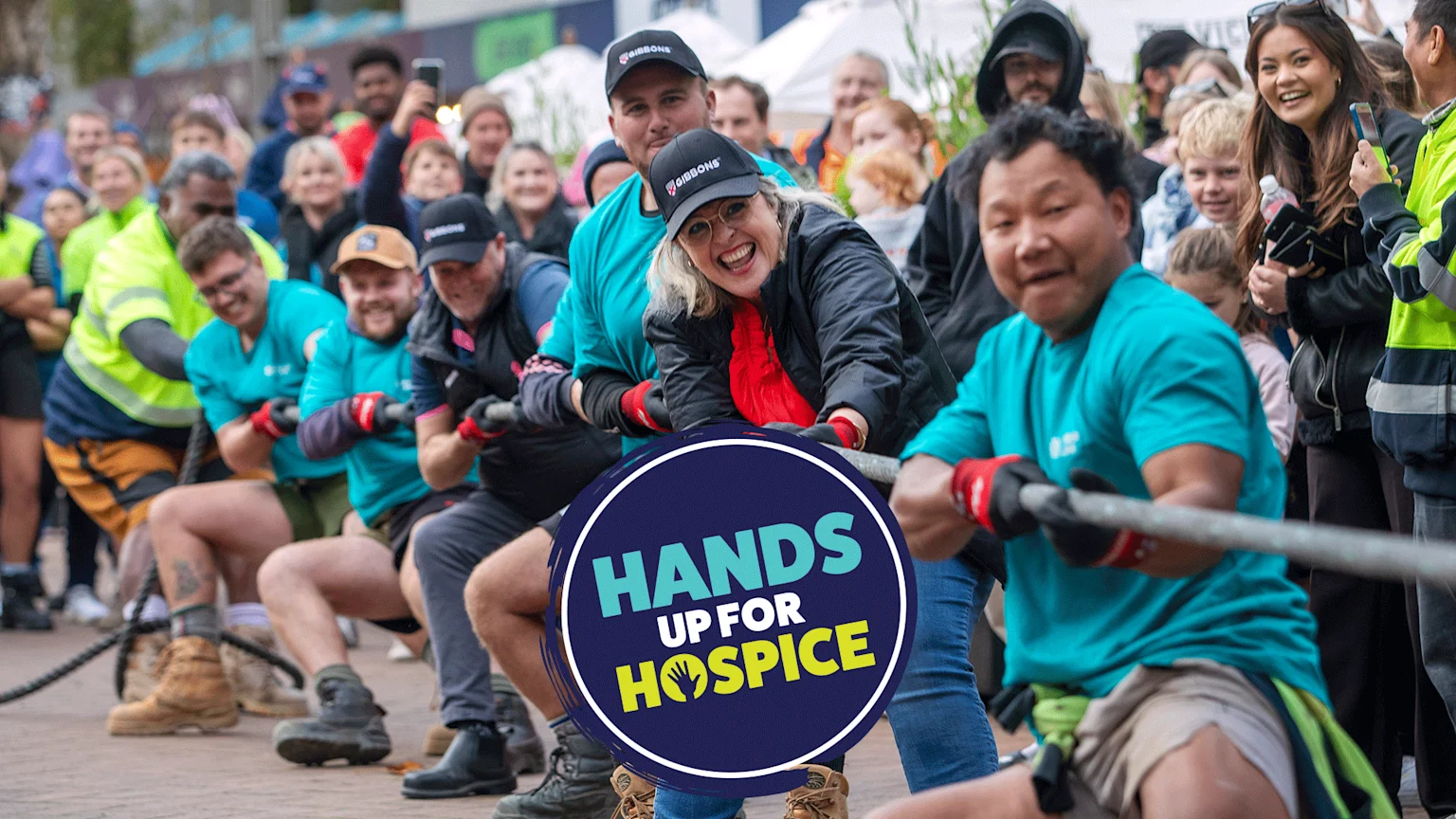 A diverse group of enthusiastic adults participates in a tug-of-war, pulling a thick rope with smiles, while a circular "Hands Up For Hospice" logo is overlaid in the center foreground.